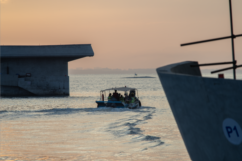 Small fishing boat navigating calm water at sunrise between moored vessels and a concrete structure.