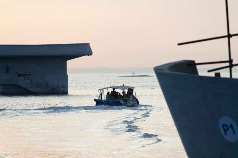 Small fishing boat moving through shallow water at dawn, framed by the hulls of larger beached vessels.