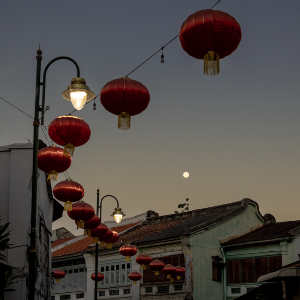 Red paper lanterns strung along a pole with traditional architecture at dusk, moon visible in sky.