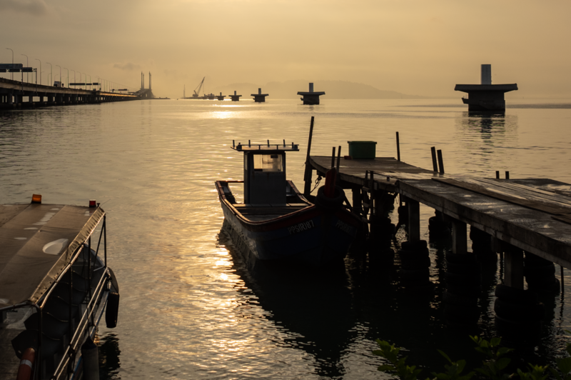 Silhouetted fishing boats moored at wooden docks during golden hour, with additional vessels visible in the misty background across calm water.
