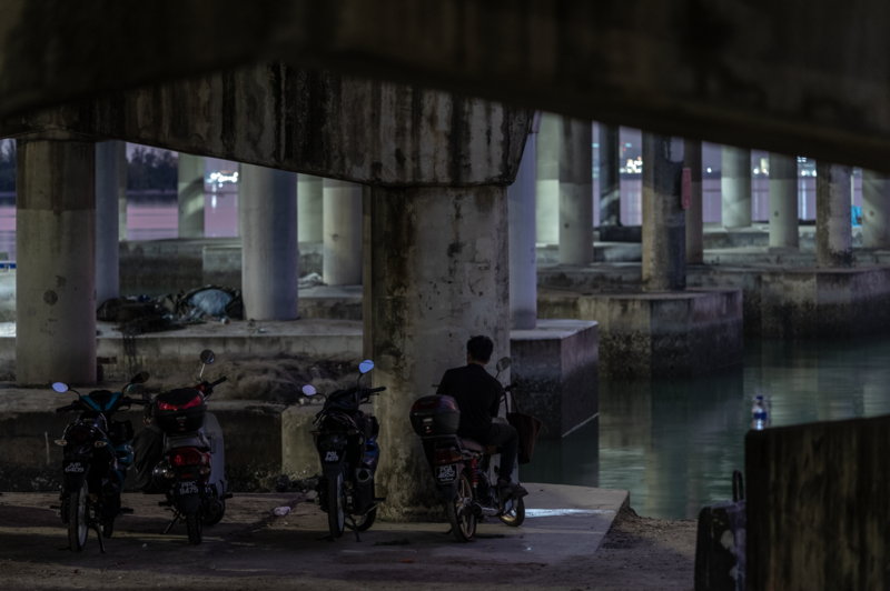 Person standing with motorcycles beneath an underpass at dusk, with water and lit buildings visible in background.