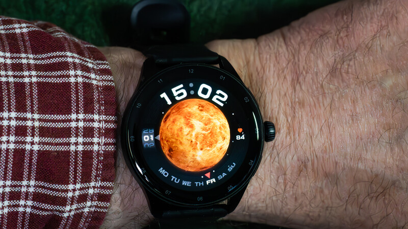 Smartwatch displaying time 15:02 with glowing sun illustration on black dial against plaid fabric background.