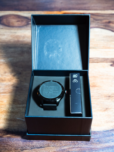 Smartwatch with black band and charging dock displayed in open luxury box on wooden surface.
