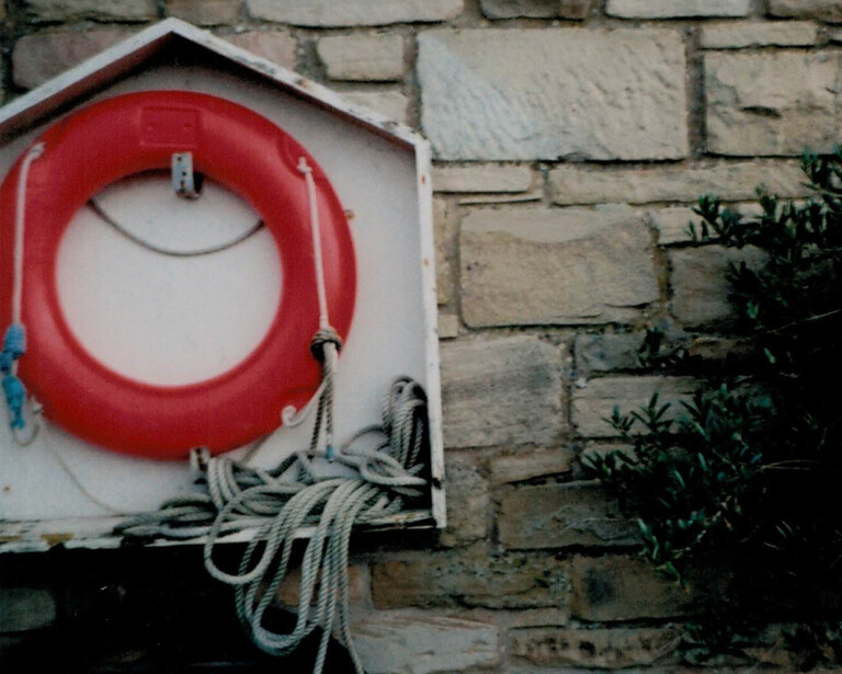 Red life ring mounted on white panel against weathered stone wall with coiled white rope beneath.