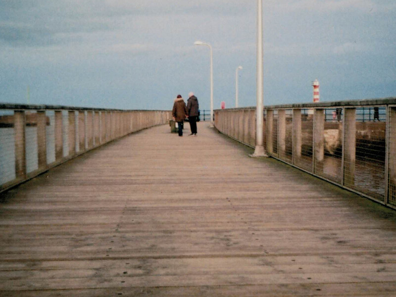 Two figures walking away on a wooden pier with metal railings extending toward a distant lighthouse.