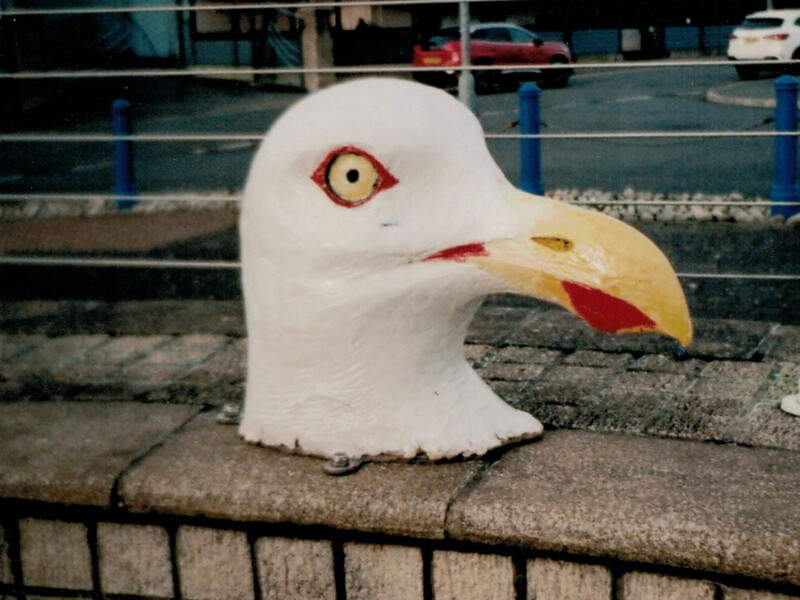 Close-up of a white bird head sculpture with yellow beak and red eye detail displayed on concrete ledge.