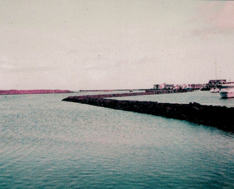 Curved dark breakwater extending into turquoise shallow water with pink sky and distant buildings.