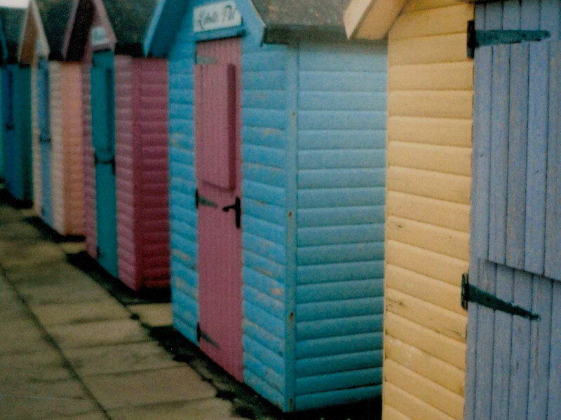 Row of colorful beach huts in turquoise, pink, and cream with gray doors along a concrete promenade.