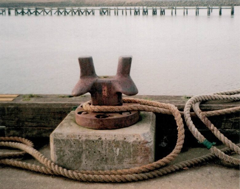 Weathered metal bollard with rope coiled around its base on a concrete dock.