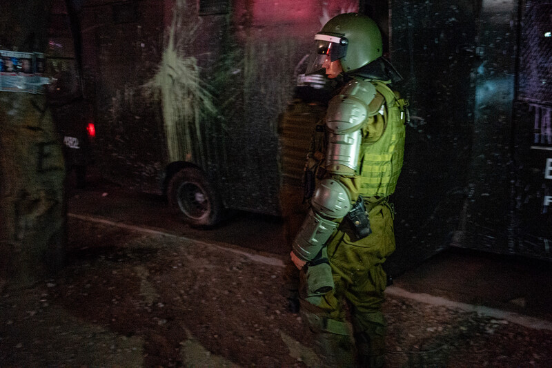 Night scene with military personnel in tactical gear illuminated by vehicle headlights in an urban alley.