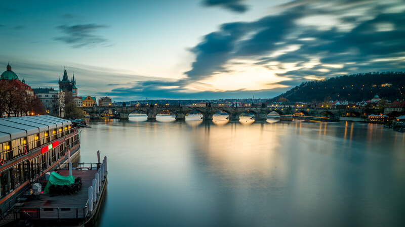 Medieval stone bridge spanning a river at dusk, with illuminated buildings and church spires reflected in still water.