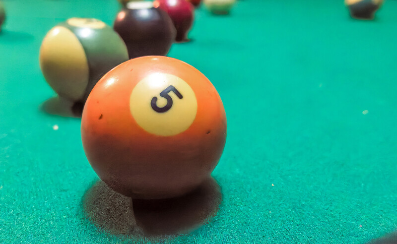 Orange billiard ball numbered 5 in sharp focus on turquoise pool table felt, with additional balls blurred in background.