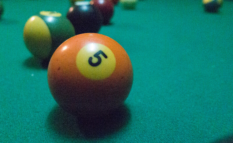 Orange billiard ball numbered 5 in sharp focus on turquoise pool table felt with other balls blurred in background.