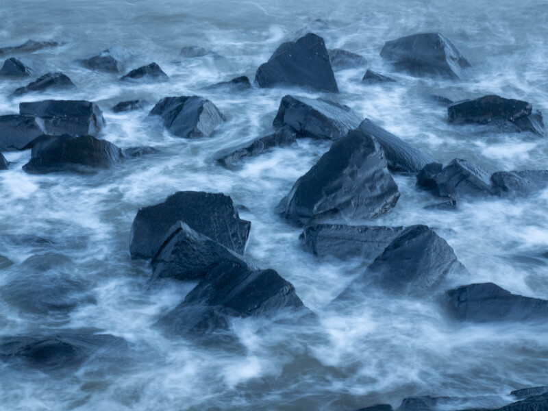 Waves crash around dark rocks jutting from the ocean surface, creating white foam and spray.