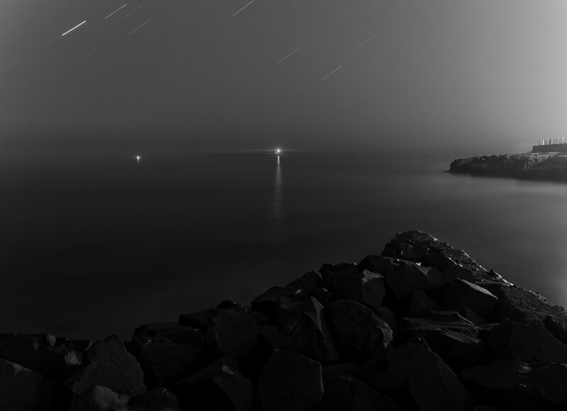Long exposure photograph of a rocky jetty extending into still water under overcast sky with distant lights.