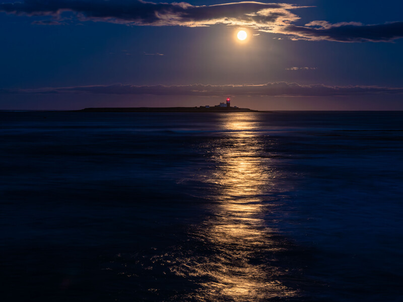 Full moon illuminating a golden path across dark ocean water toward a distant lighthouse on the horizon.