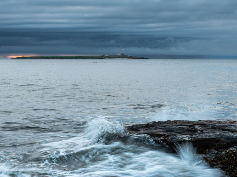Waves crash against dark rocks on a beach at dusk, with a distant lighthouse on the horizon under stormy skies.