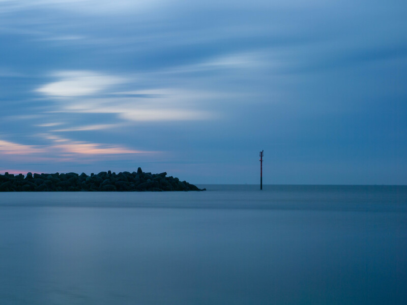 Calm seascape at dusk with distant headland and solitary post in still water under blue sky.