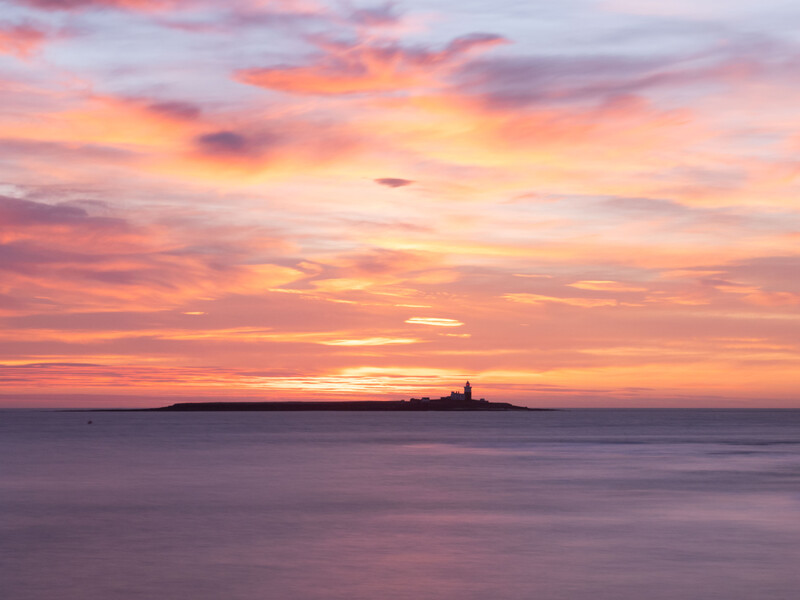 Silhouetted lighthouse on a distant island at sunset with golden and pink sky reflected in calm water.