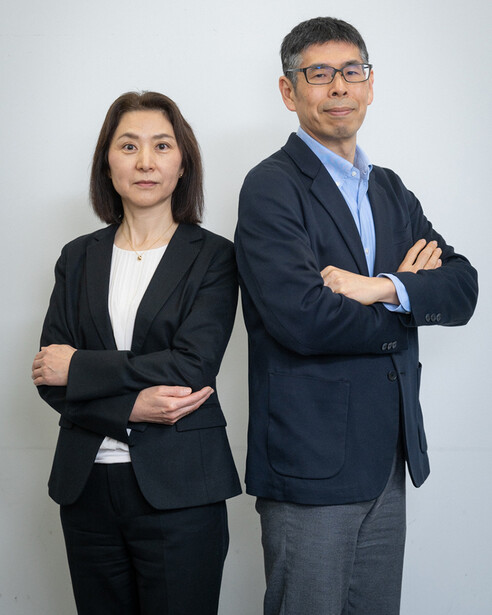 Two business professionals in dark blazers stand back-to-back with arms crossed against a white background.