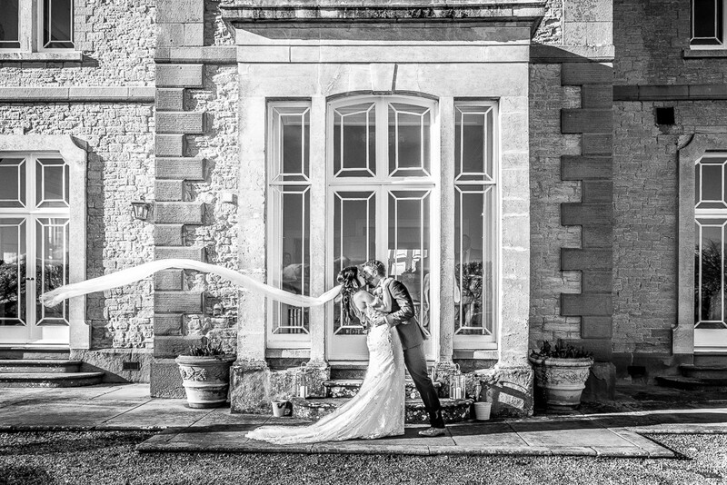Bride and groom embracing in front of a grand stone mansion with tall arched windows, photographed in black and white.