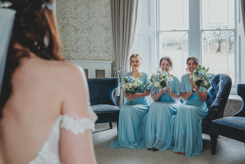 Bride in white dress viewing three bridesmaids in blue gowns holding white flower bouquets seated by large windows.