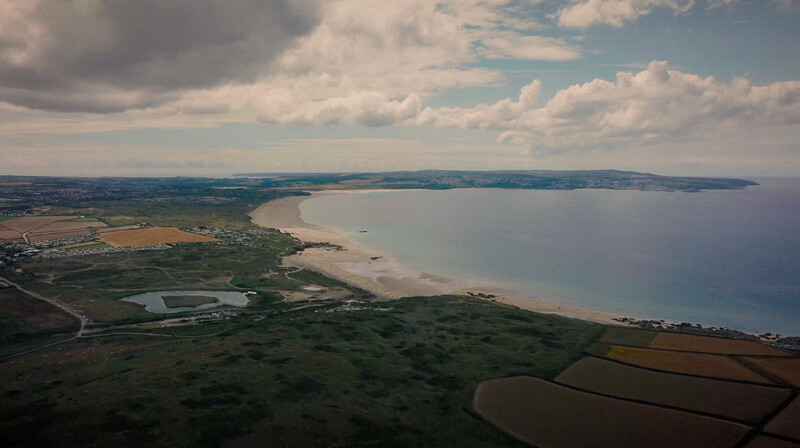 Aerial view of a coastal landscape with sandy beach, green fields, and calm waters under partly cloudy sky.
