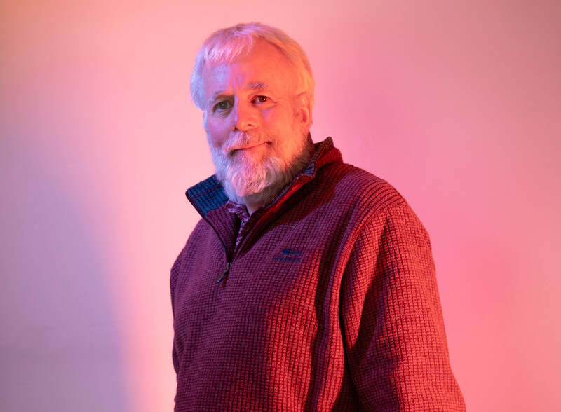 Older man with white beard wearing a red textured sweater against a pink and purple gradient background.