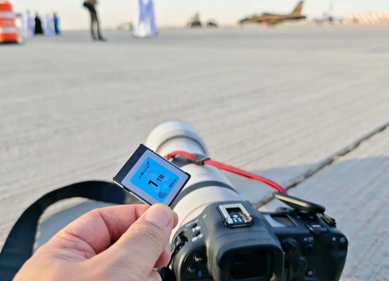 Hand holding a light meter displaying a blue screen with reading information against a beach backdrop.