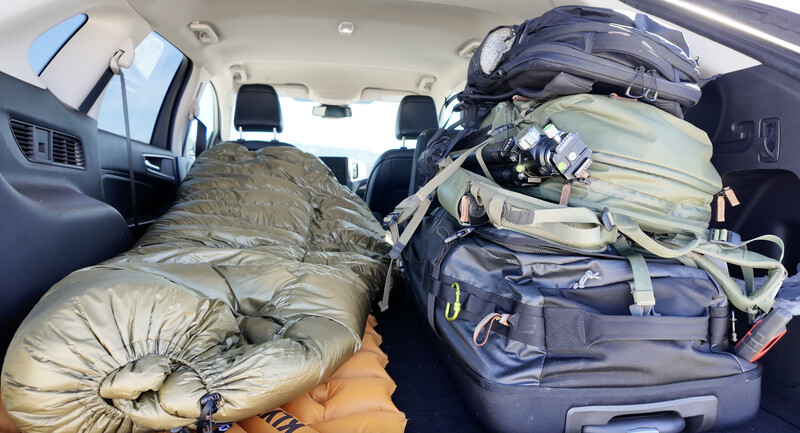 Van interior packed with sleeping bags, duffel bags, and various travel gear stacked on seats and floor.