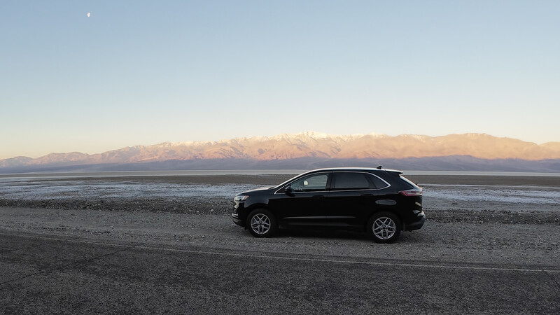 Black SUV parked on a beach with snow-capped mountains across the water at dawn or dusk.