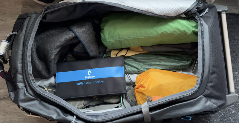 Open travel bag packed with outdoor gear including a solar charger, sleeping bag, and clothing layers.