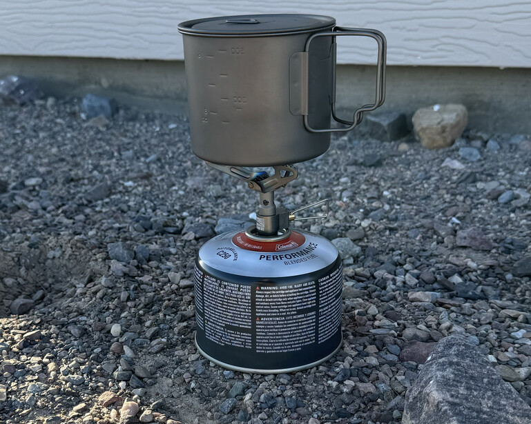 Portable camping stove with fuel canister on gravelly beach near water.