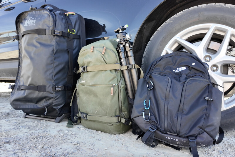 Three camera backpacks of varying sizes lined up beside an RV wheel on gravel.