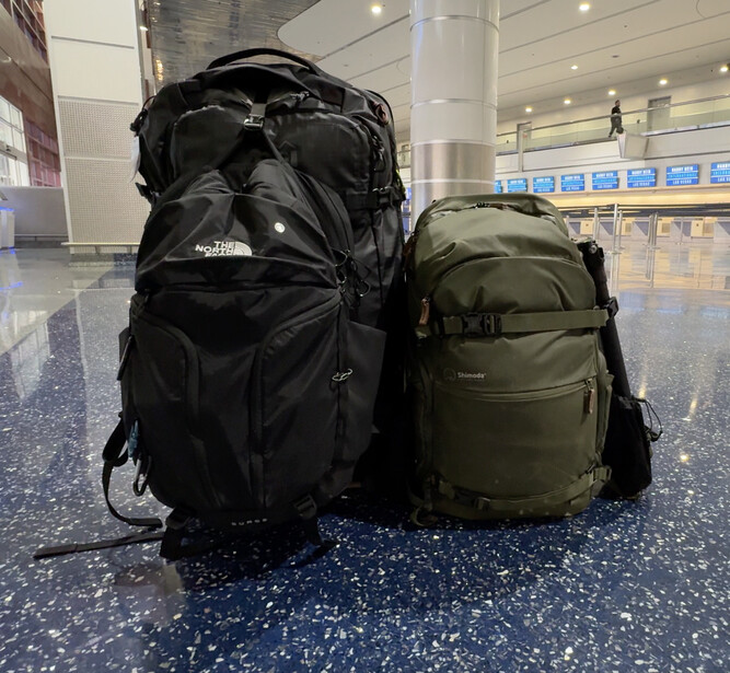 Two hiking backpacks sitting side-by-side on a polished airport floor, one black and one olive green.