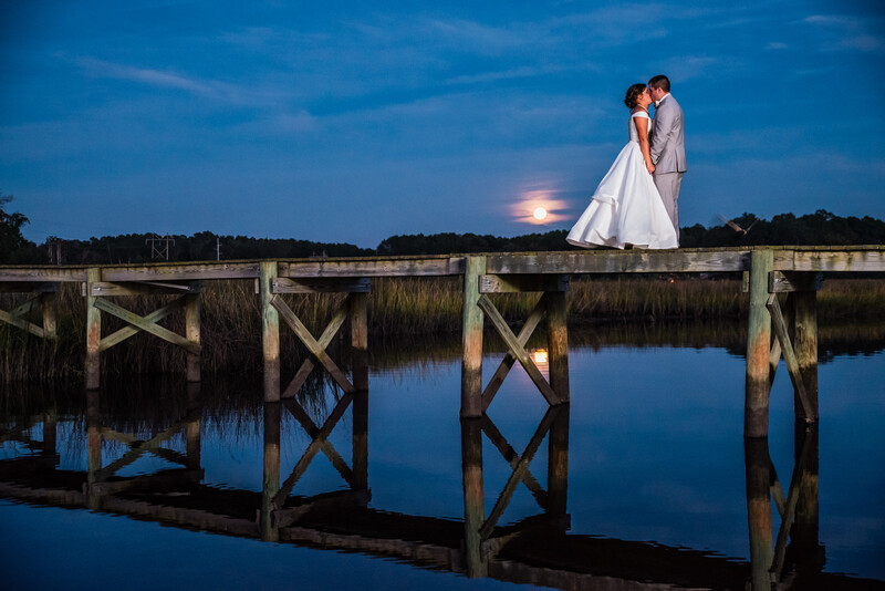 Couple in wedding attire embracing on a wooden pier at twilight with full moon rising over calm water.