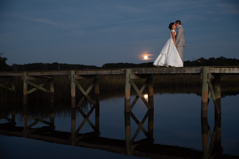 Bride and groom kissing on a wooden pier at twilight with a full moon rising over water.