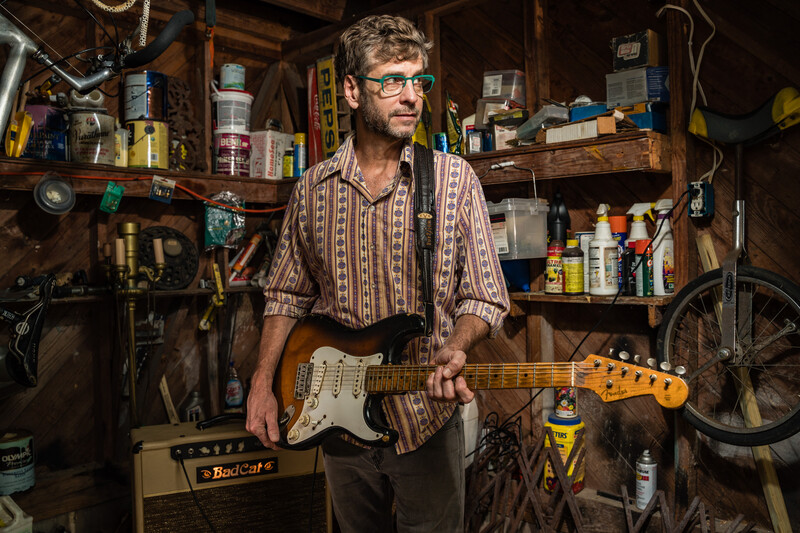 Musician holding a Fender Stratocaster in a cluttered workshop filled with tools, electronics, and vintage equipment.