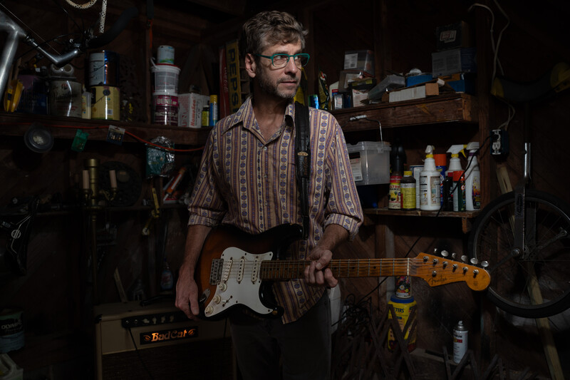 Musician holding a Fender Stratocaster guitar in a workshop studio setting.