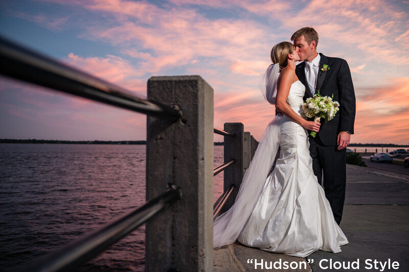 Bride and groom kissing on a waterfront dock at sunset with a bridge visible in the background.