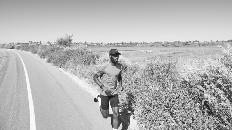 Runner jogging along a straight rural road lined with scrubland vegetation.