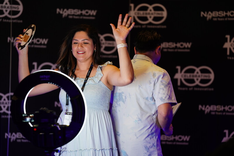 Woman in light blue dress waving at camera during event, with ring light visible in foreground.