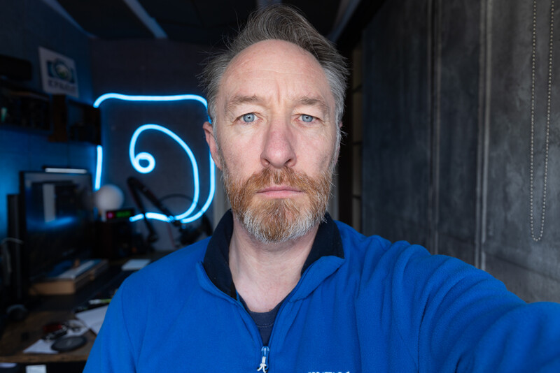Man with beard wearing blue jacket in front of neon blue sign in studio setting.