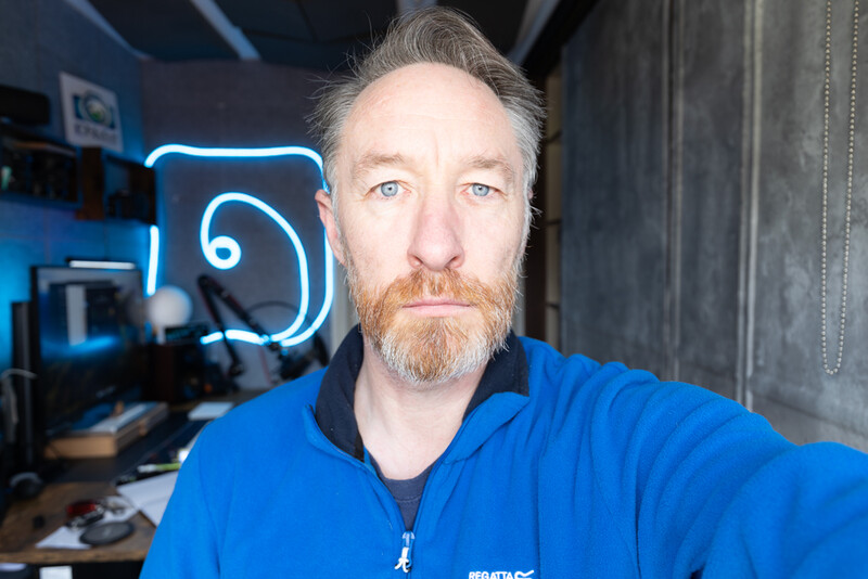 Man in blue jacket with beard posing in front of neon sign in studio setting.