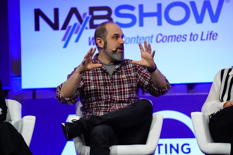 Man speaking animatedly at a conference panel, gesturing with both hands while seated on a white chair.