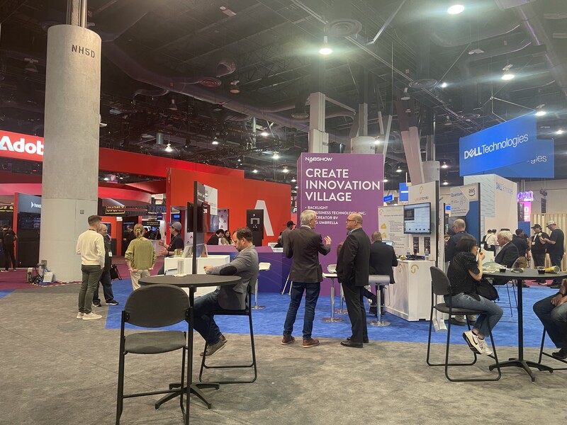Interior of a large convention center or trade show with red and purple branded booths, display walls, and attendees networking at tables under industrial ceiling lights.
