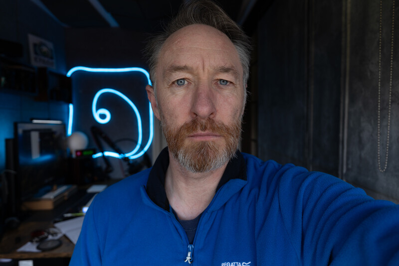 Man in blue jacket photographed indoors with neon sign in background.