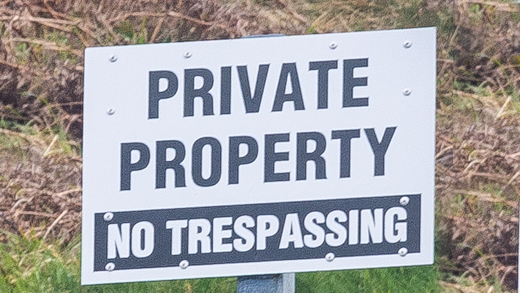 White sign with black text reading 'PRIVATE PROPERTY' and 'NO TRESPASSING' mounted on a post in dry grass.