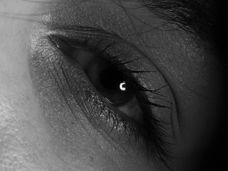 Close-up black and white portrait of an eye with defined eyeliner and long lashes, with a crescent moon reflection visible in the pupil.
