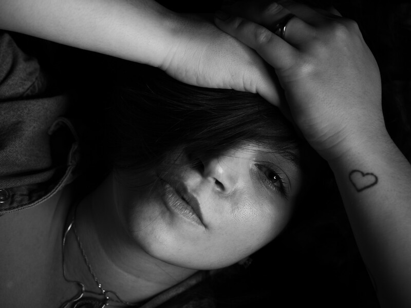 Black and white portrait of a young person with hand raised to head, displaying a small heart tattoo on their arm.